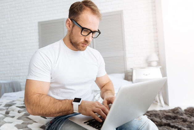 技术我的自由职业者魅力项目专注的大胡子男人坐在家里的床上 一边用笔记本电脑一边表达专注和工作积极性白天创新
