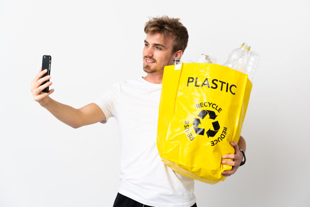 志愿者年轻的金发男人拿着一个装满纸的回收袋 在白色的垃圾桶里自拍青年瓶子垃圾
