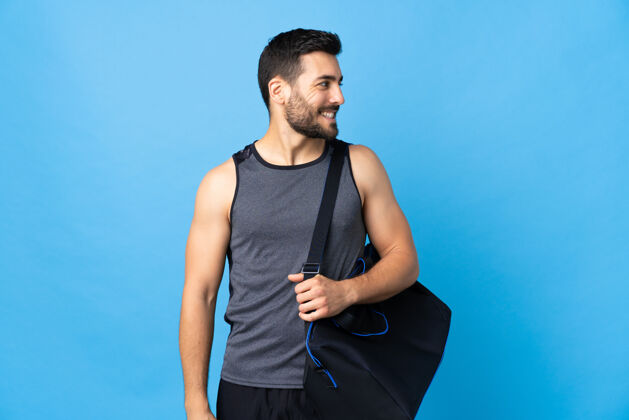 手势年轻的运动男子与运动包孤立的蓝色背景看一边健身房胡须看