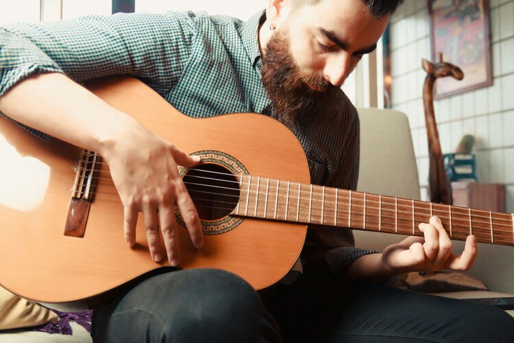 表达一个留着胡须和衬衫的年轻潮人 在晴朗的日子里在沙发上演奏西班牙吉他西班牙语音乐学习
