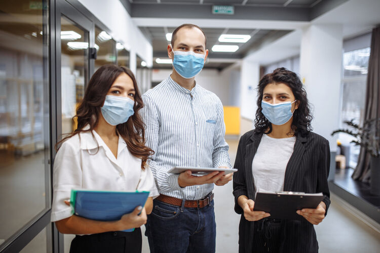 商业女人三个戴着医用口罩的同事在冠状病毒大流行隔离期间在办公室走廊讨论业务团队文件男人