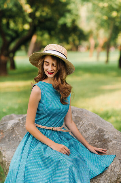 服装一个穿着蓝色裙子 戴着一顶优雅帽子的时髦女孩坐在公园里的一块大石头上的画像石头花园室内