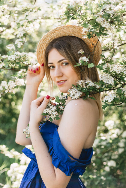 年轻美丽的时髦女人在盛开的灌木中的感性平静的肖像 白色的花朵绣线菊空间时尚金发碧眼的女人在花园里穿着蓝裙子戴着蓝帽子时尚欢呼户外