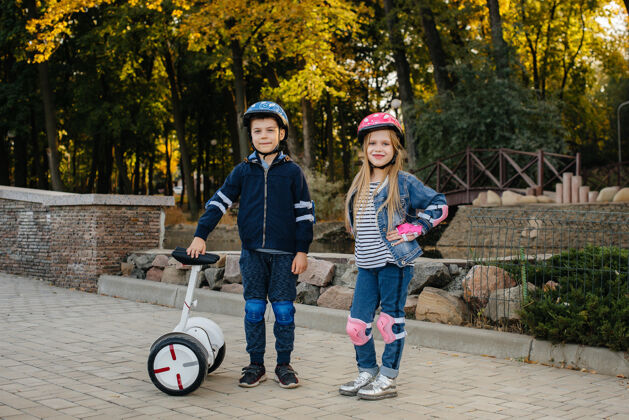 男孩快乐的男孩和女孩站在公园骑赛格威和滑板车在日落家庭头盔设备