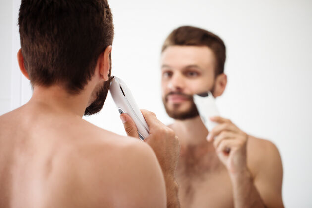 修剪英俊的年轻人在街上刮胡子浴室.肖像一个时髦的裸体胡须男人在家里的镜子里检查他的脸治疗男人护理