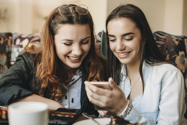 人两位可爱的女性朋友在咖啡馆玩得很开心 一边看着智能手机笑着的照片影响者电话坐着