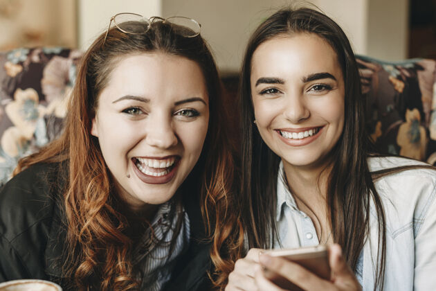 饮食两个可爱的女性坐在咖啡馆里拿着智能手机笑着看着相机的特写镜头红发桌子朋友
