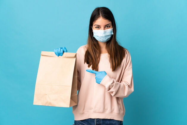 健康一位年轻的女士戴着口罩 拿着一个隔离在蓝色背景上的购物袋 指着它 保护自己免受冠状病毒的侵害面具表情指着