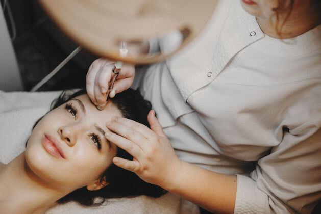 痤疮俯视图：美丽的女性脸靠在水疗床上 睁开眼睛 在水疗中心的美容师对她的脸进行无创性的微晶磨皮术医学人美容