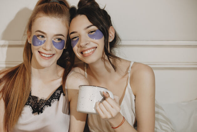 朋友两位可爱的年轻女性一边做面部护理一边喝着热茶 劳金躺在床上休息的照片化妆品年轻面部