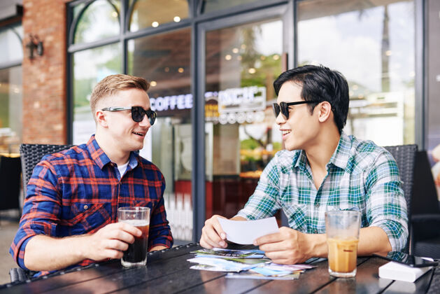 亚洲人笑容可掬的年轻帅哥戴着墨镜坐在咖啡桌旁 讨论着夏日旅行的打印照片男人微笑讨论