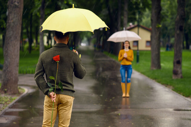拥抱雨中公园浪漫约会戴曼带着红玫瑰在小路上等着他的女人 小巷里天气潮湿夏天关系一起