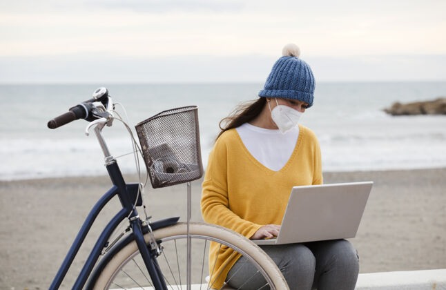 海洋一位戴着口罩的数字营销人员正坐在木板路上与一位女士一起做一个新的笔记本电脑推广项目自行车独立女性与客户沟通时尚休闲海滩