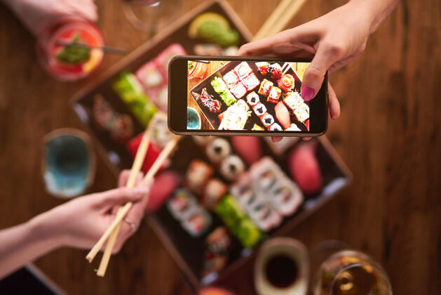 酱油顶部视图2女孩们正在吃寿司和拍照智能手机.set各种面包卷和寿司配饮料亚洲菜肴美味