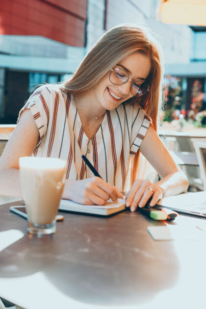 杯子可爱的年轻女性 满头红发 满脸雀斑 一边在户外一边喝咖啡一边在笔记本上写字眼镜连接咖啡