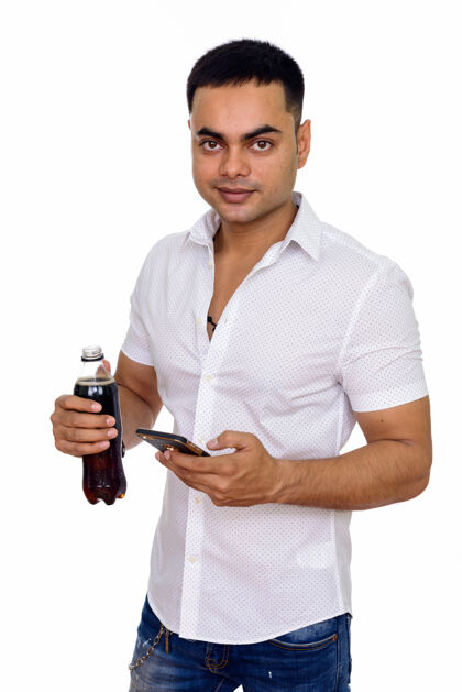持有年轻英俊的印度男人一边用手机一边拿着一瓶苏打水互联网信息饮食