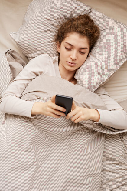 抱早上在床上使用手机的年轻女子的高角度视图枕头触摸屏家居