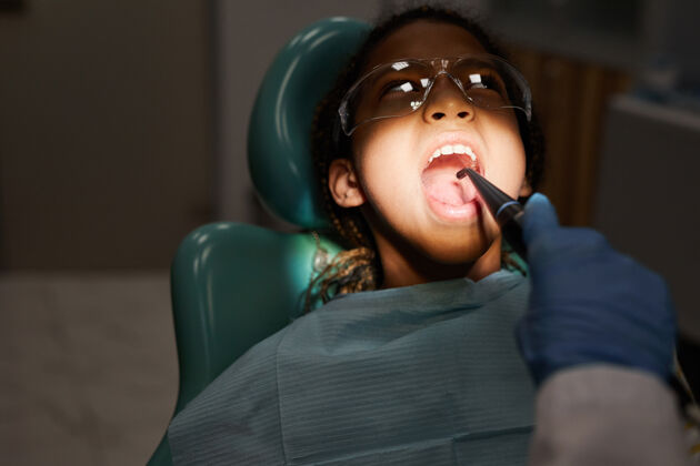 室内戴着护目镜的小病人在牙医用牙钻治疗牙齿时张开嘴医生美丽女孩