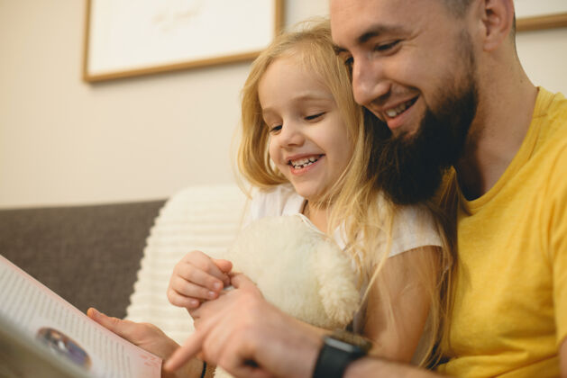 可爱一个可爱的小女孩和她年轻的留胡子的父亲早上在家里的沙发上看书的画像女儿胡子年轻
