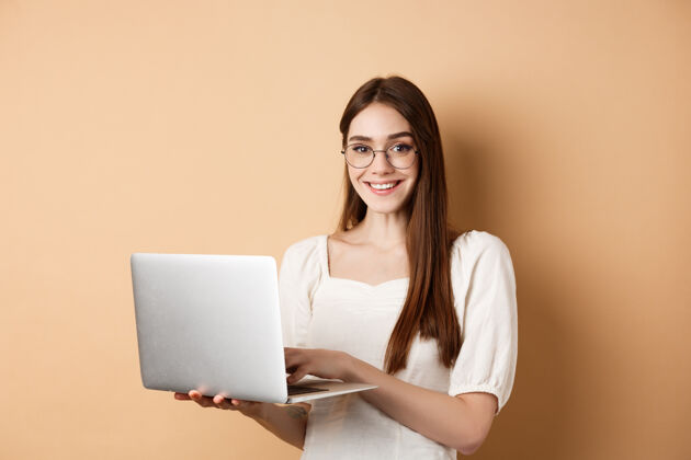 笔记本电脑面带微笑的女学生在笔记本电脑上工作 戴着眼镜 看起来很开心 站在米色的背景下使用电脑自然候选人化妆品
