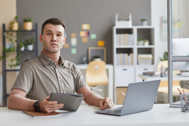 互联网年轻的平面设计师坐在桌子前看着面前的肖像他在工作中使用数字平板电脑和笔记本电脑职业技术肖像