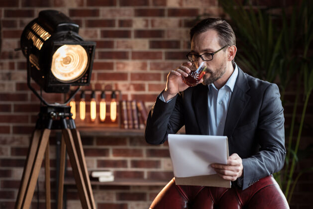 男士昂贵的酒精帅哥严肃时尚的男人站在扶手椅旁 一边喝着威士忌一边读笔记白天现代玻璃