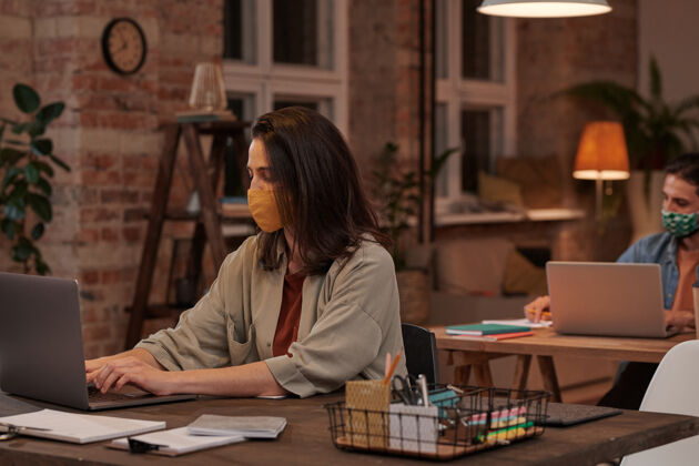 休闲服戴着防护面具的年轻女商人坐在桌子旁 晚上在办公室用笔记本电脑打字商务女性看着流行
