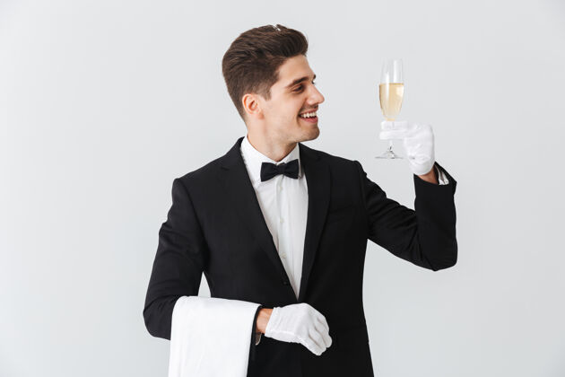 人身着燕尾服的微笑年轻侍者的画像在白墙上为您奉上一杯香槟信心工作杯子
