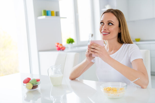 年轻梦想家主妇拿着热饮料早餐玉米片健康饮食看着梦幻想象自己下巴坐在桌子白光厨房室内牛奶杯子咖啡