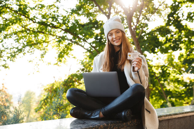肖像快乐的年轻女子穿着秋大衣 戴着秋帽坐在户外 用笔记本电脑 展示塑料信用卡概念电脑街道