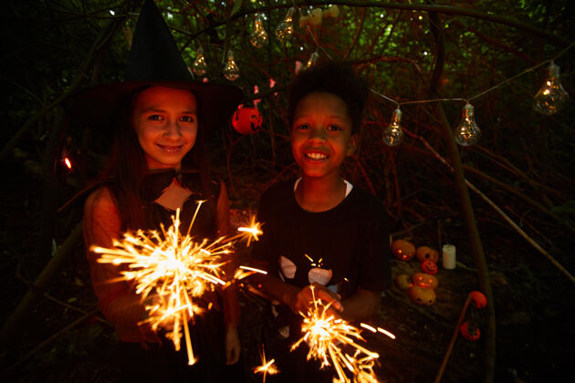 享受照片中的朋友们手持火花 对着镜头微笑 站在黑暗的森林里 一起庆祝万圣节季节夜晚火花