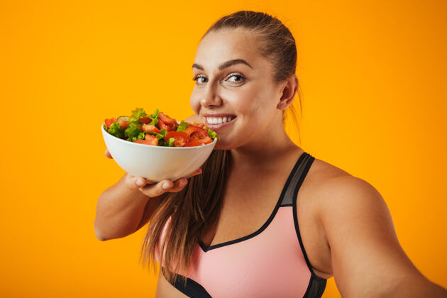 体重一个穿着运动服的快乐的超重健身妇女的肖像 孤立地站在黄色的墙上 用手机自拍 展示盛着沙拉的碗身体午餐健身
