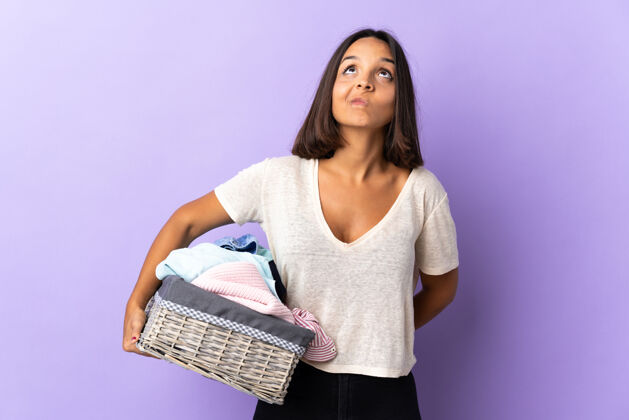 看年轻的拉丁女子拿着一个衣篮孤立地站在紫色地上仰望洗衣人脏
