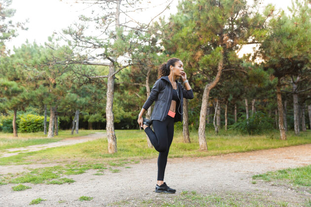 私人教练一个20多岁的非裔美国妇女穿着黑色运动服 在绿色公园里伸展身体锻炼音乐身体