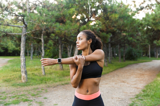 听力年轻的非洲裔美国妇女20多岁穿着黑色运动服做运动 并伸展她的身体在绿色公园无线运动手臂