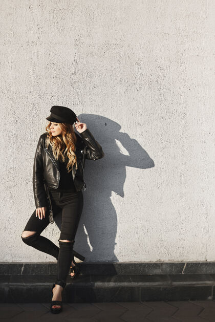 年轻穿着皮夹克 破牛仔裤 戴着黑帽子的时髦金发模特儿站在户外 靠着城市的城墙帽子衣服皮革