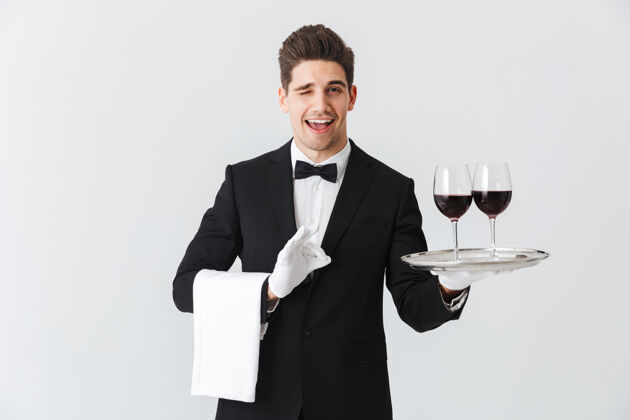 夹克穿着燕尾服的年轻帅哥侍者端着一个盘子 灰色的墙上隔着两杯红酒酒正式管家