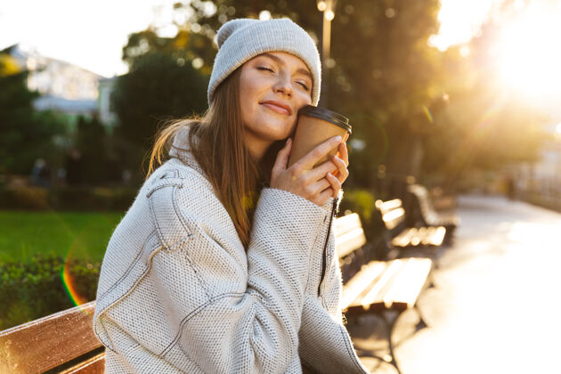 城市美丽的年轻女子穿着秋大衣 戴着秋帽 坐在户外的长凳上 端着一杯热咖啡享受长凳休闲