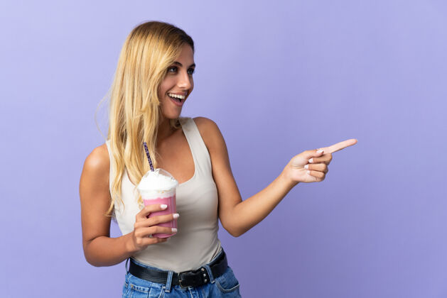 年轻人年轻的金发乌拉圭女人用紫色的手指指着旁边 拿着草莓奶昔 展示一款产品草莓奶昔人