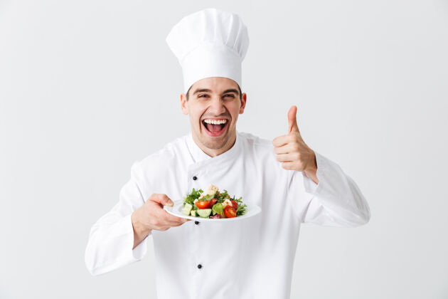 准备兴奋的男厨师穿着制服 把新鲜的蔬菜沙拉放在隔离在白色墙上的盘子上厨师烹饪信心