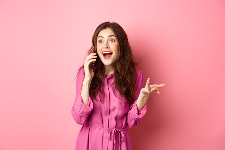 应用程序快乐的黑发女人在手机上聊天 做手势和笑 随意的有趣的交谈 站在粉红色的墙上乐观时尚咧嘴笑
