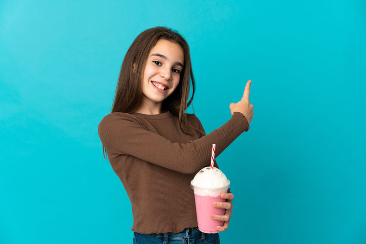 手指小女孩拿着草莓奶昔孤立在蓝色的墙上指着后面回来杯子食物