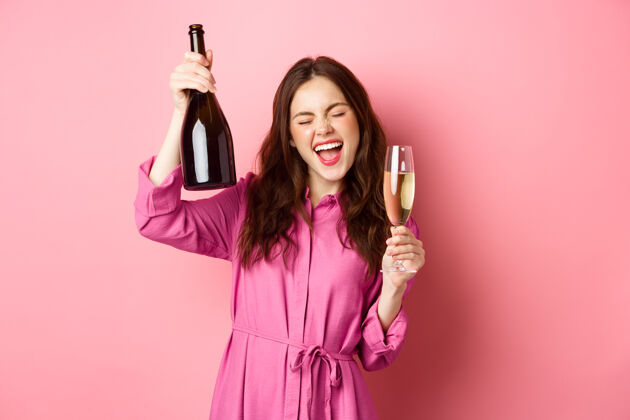 积极庆祝和节日的概念快乐的女人跳舞和娱乐 喝香槟 拿着酒瓶和玻璃杯 烤面包 站在粉红色的墙上复制空间请候选人兴奋