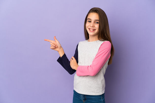 快乐小女孩孤立地站在紫色的墙上 手指指向一边眼睛欢迎童年