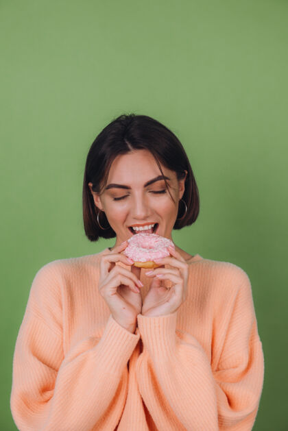 可爱穿着休闲桃色毛衣的年轻时尚女性 隔离在绿色橄榄色墙壁上 粉色甜甜圈快乐复制空间甜点釉面漂亮