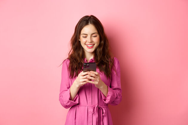 手机年轻女子购物 手机聊天 微笑着看智能手机屏幕 站在粉红色的墙上复制空间手势赞异科技