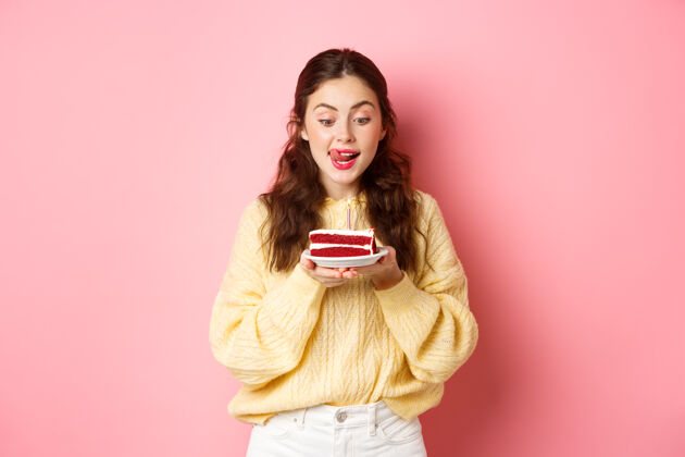 休闲庆祝和节日生日快乐的女孩舔着嘴唇 带着诱惑的表情看着生日蛋糕 想咬一口 站在粉红色的墙上时尚蛋糕生日