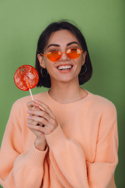 魅力年轻时尚的女士穿着休闲的桃色毛衣 戴着橙色的眼镜 隔离在绿色橄榄色的墙上 带着橙色棒棒糖的正面微笑复制空间时尚衣服成人