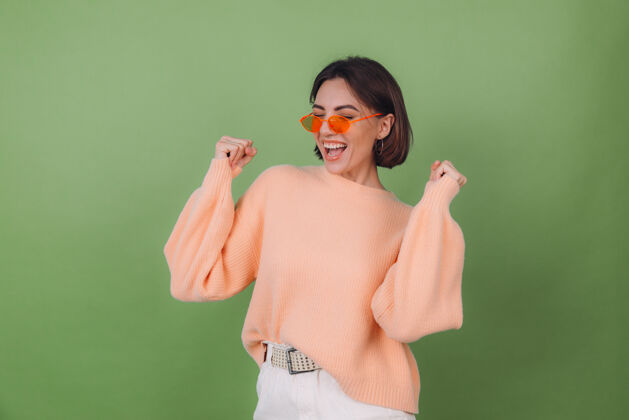 微笑年轻时尚的女士穿着休闲的桃色毛衣 戴着橙色眼镜 隔着绿橄榄色的墙壁积极地做着赢家的手势握紧拳头复制空间女性肖像人物