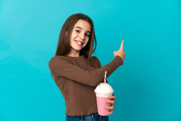 手指小女孩拿着草莓奶昔孤立在蓝色的墙上指着后面回来杯子食物
